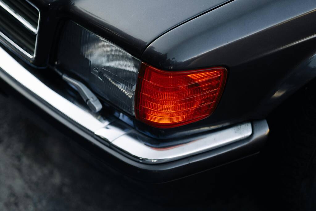 Gros plan détaillé d'un phare avant, d'un feu de position orange et d'un pare-chocs chromé d'une voiture noire classique, mettant en évidence la précision de la carrosserie et des finitions.