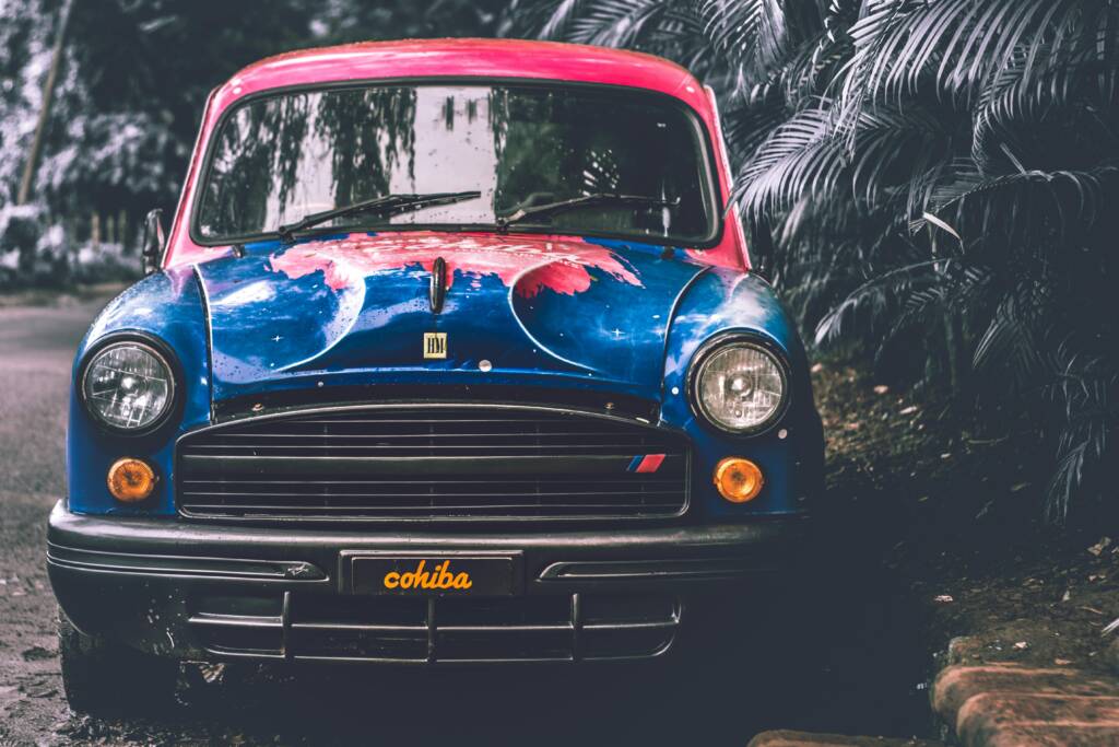Gros plan sur l'avant d'une voiture ancienne de type Hindustan Ambassador, entièrement customisée avec une peinture bleu vif, rouge et des motifs, soulignant l'art de la personnalisation.